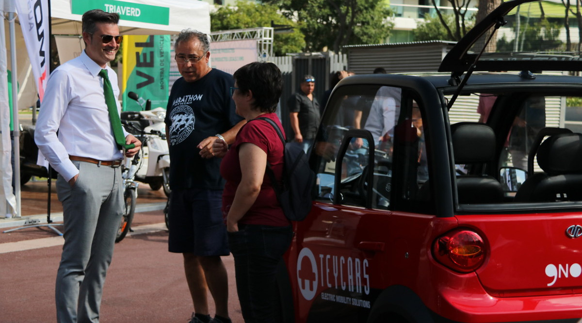 Dos visitants de Velèctric conversant amb un comercial al costat d'un cotxe elèctric. Sílvia Jardí (ACN)